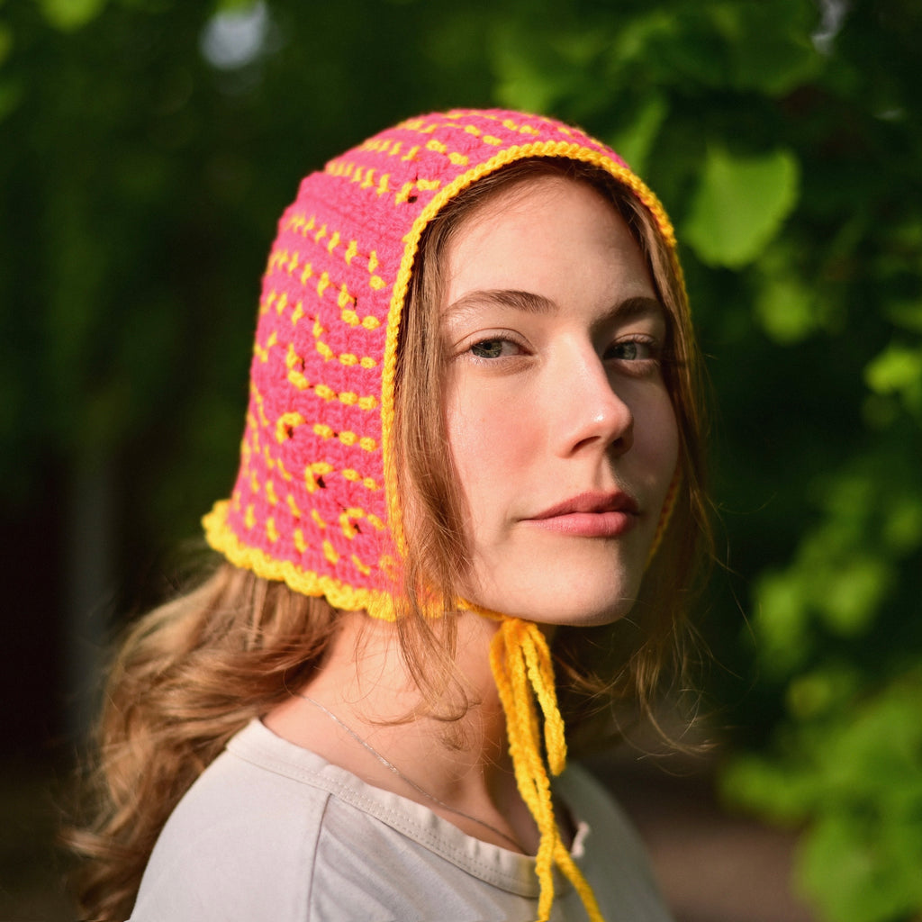Person wearing a pink and yellow crocheted bonnet outdoors with greenery in the background