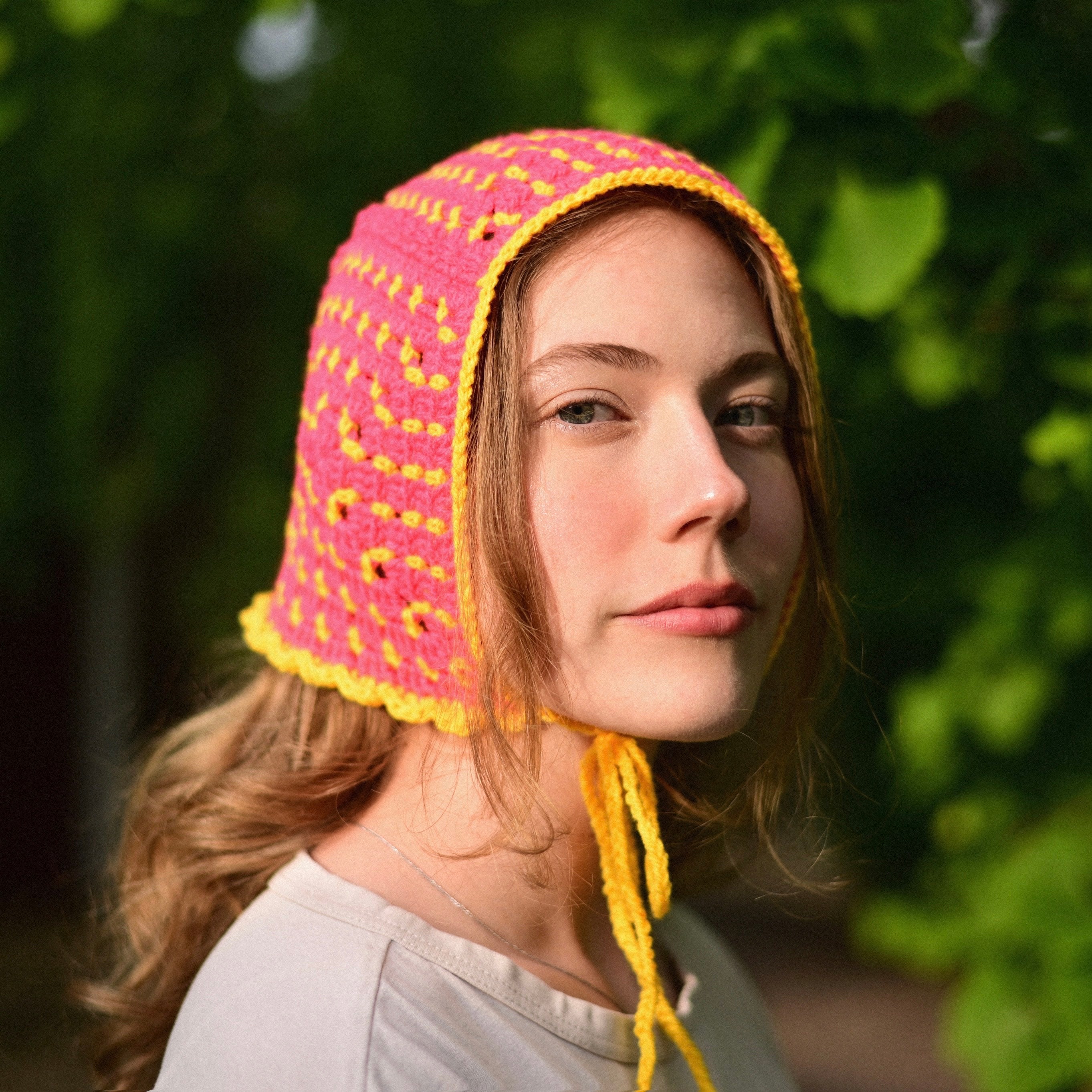 Person wearing a pink and yellow crocheted bonnet outdoors with greenery in the background