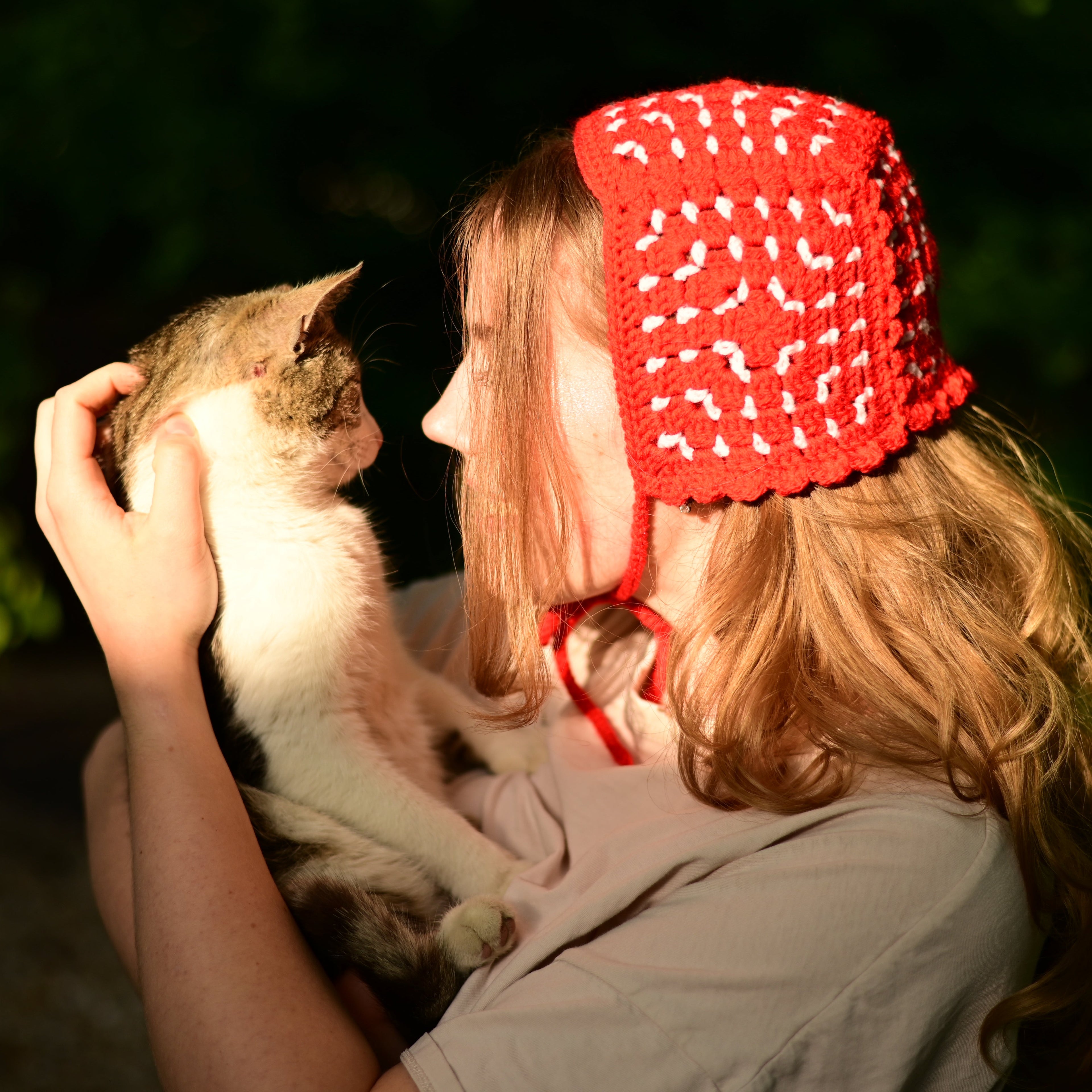 Person wearing a red crocheted bonnet holding a cat outdoors with a blurred background
