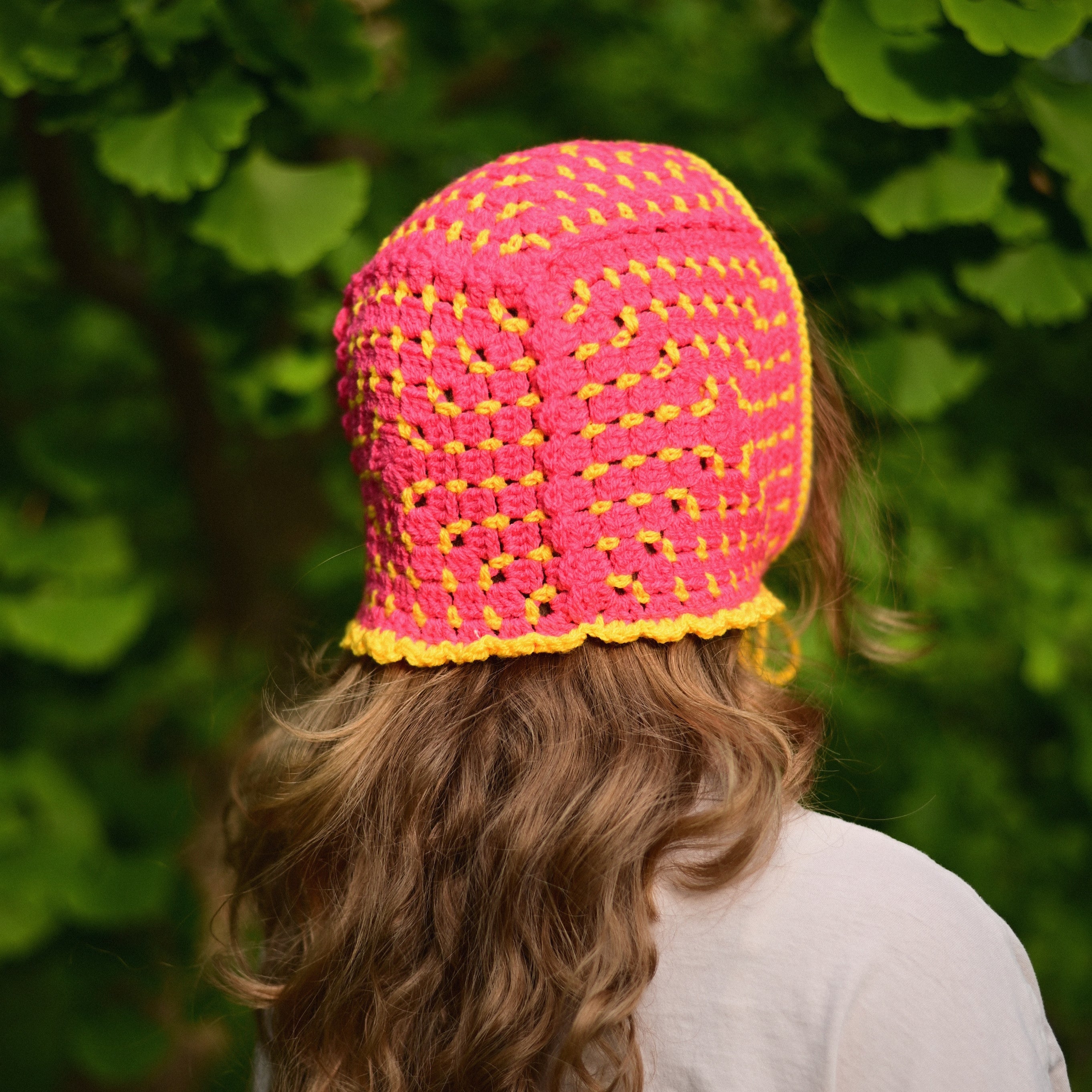 Person wearing a bright pink and yellow crocheted bonnet with a green leafy background