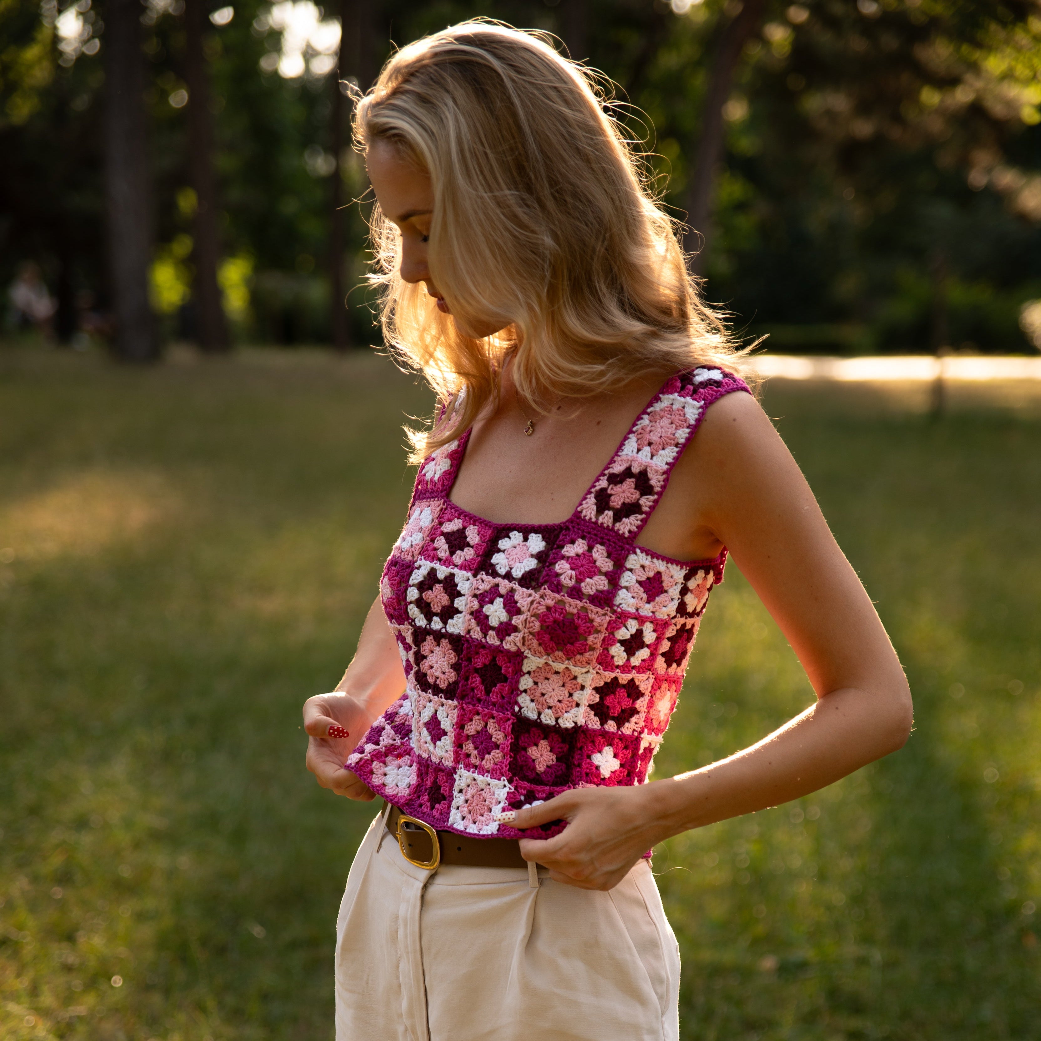 Woman wearing a colorful patchwork crochet top and beige shorts standing in a grassy area.