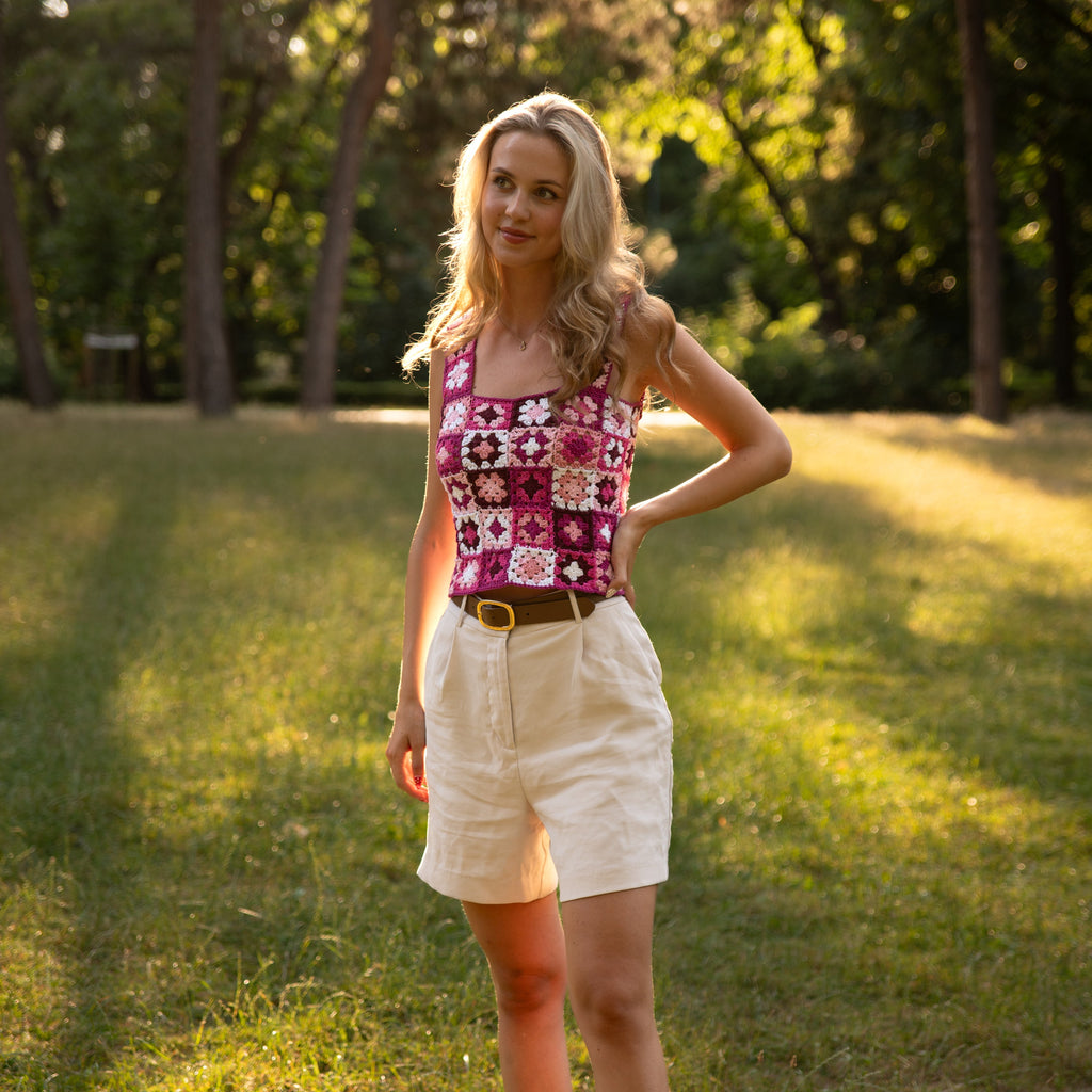 Woman in a crochet pink top and beige shorts standing in a park.