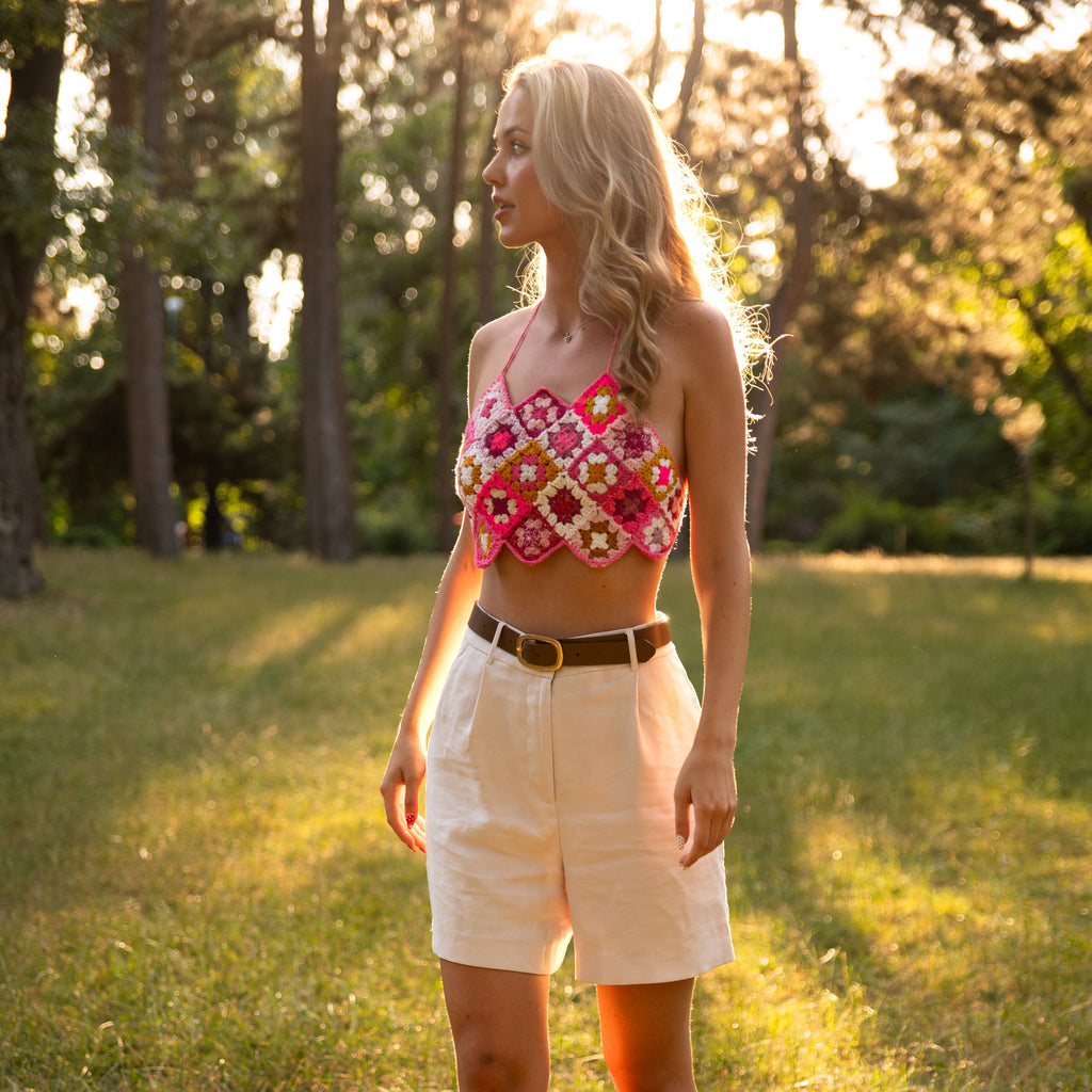 Woman wearing a pink crochet top and white shorts standing in a sunlit grassy field.