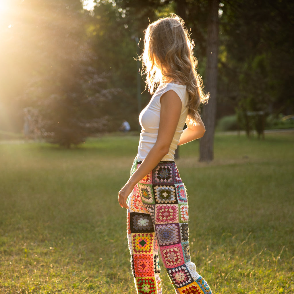 Woman wearing colorful crochet pants standing in a park with sunlight filtering through trees.