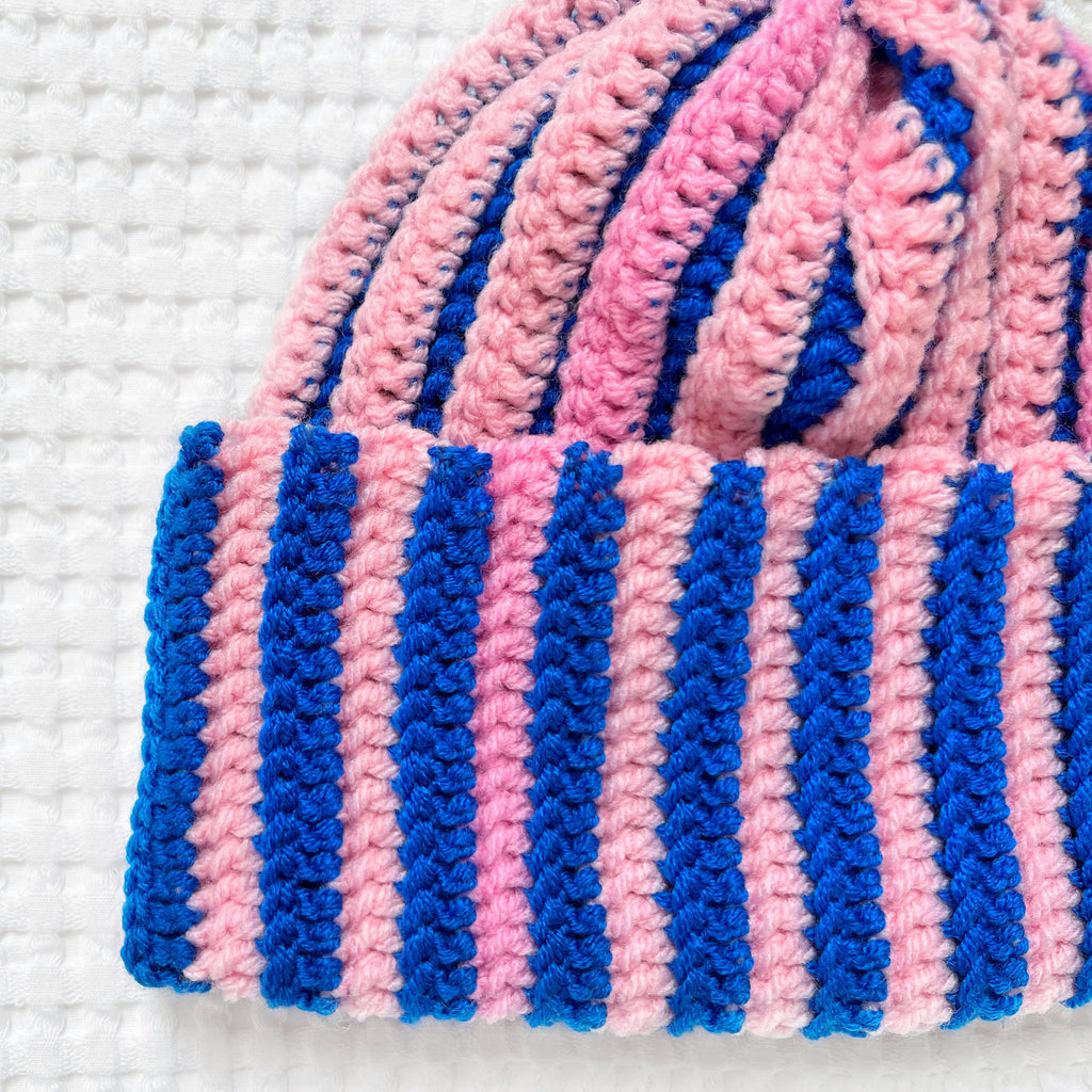 Pink and blue striped crochet beanie on a white textured background
