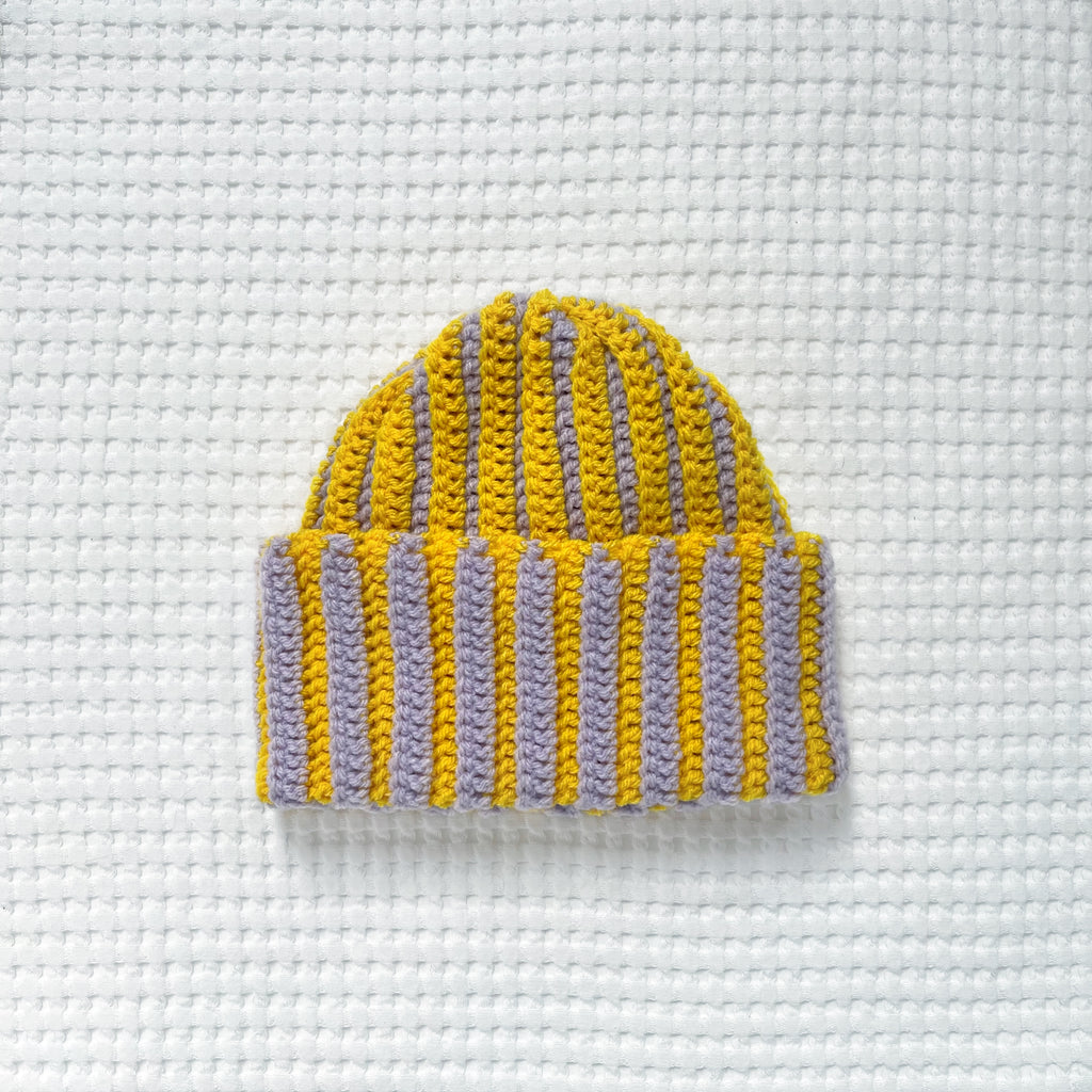 Striped yellow and pastel lilac crochet beanie on a textured white background