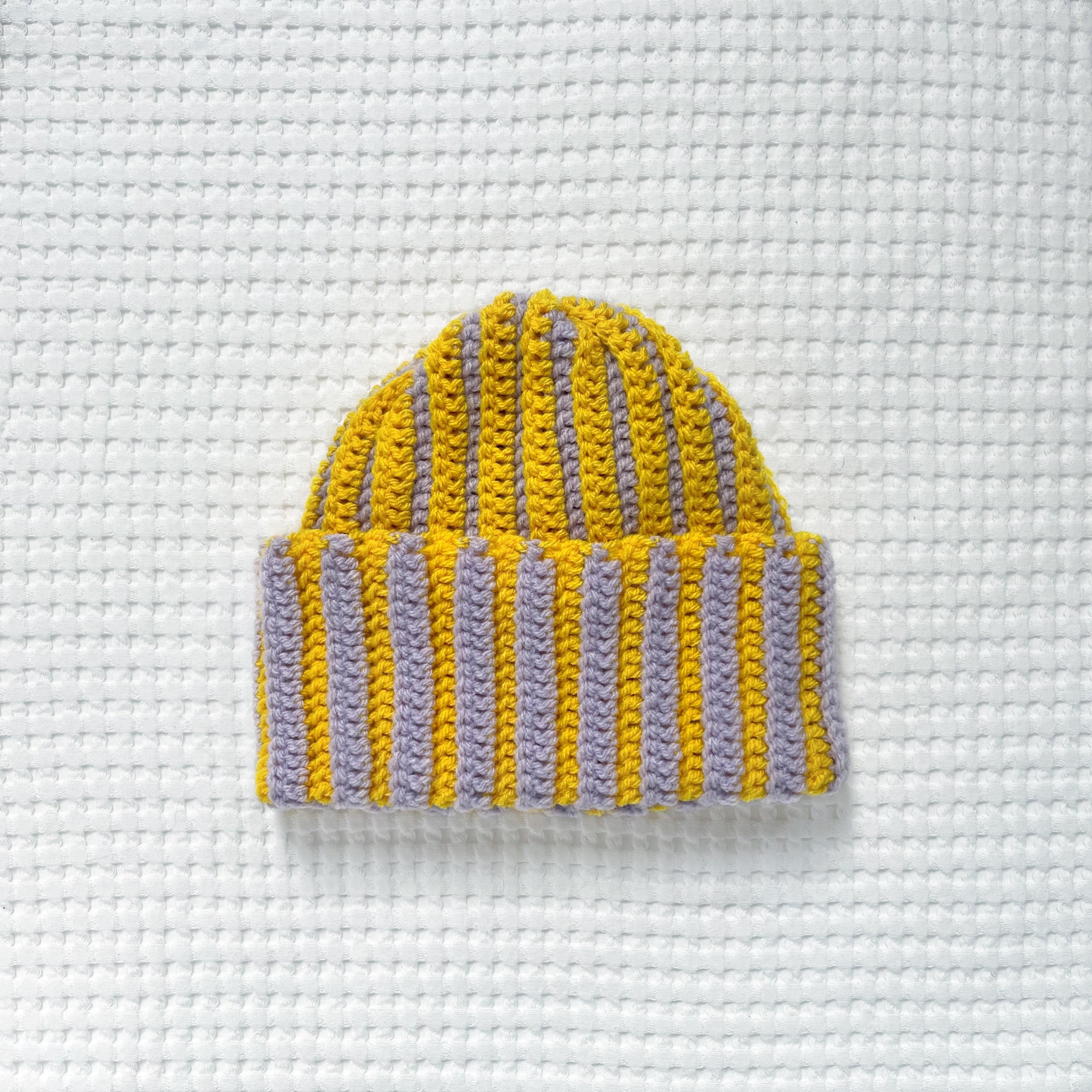 Striped yellow and pastel lilac crochet beanie on a textured white background