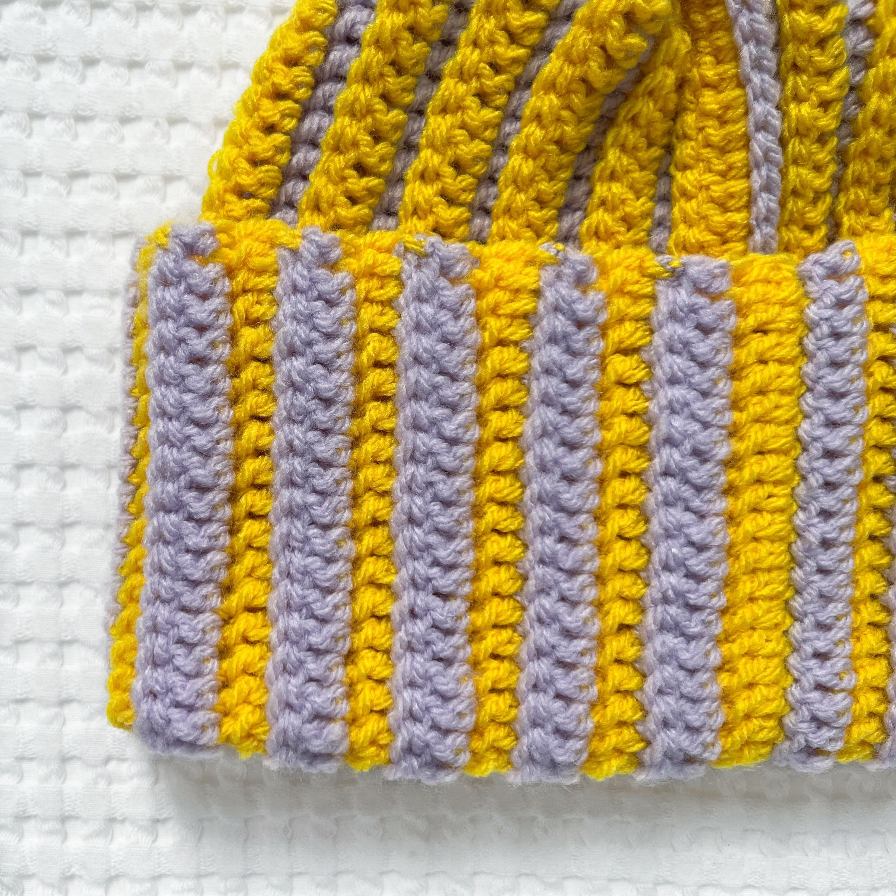 Striped yellow and pastel lilac crochet beanie on a textured white background