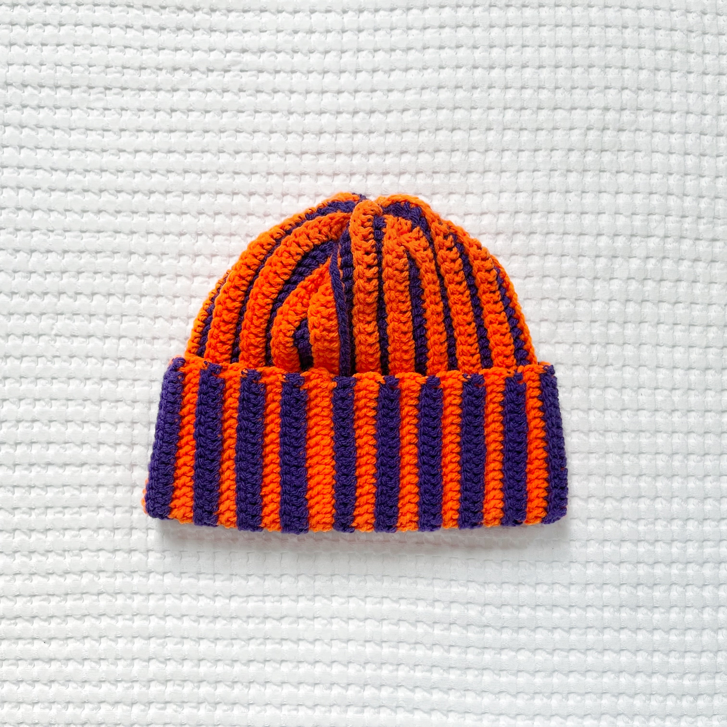 Crochet beanie with orange and purple stripes on a textured white background