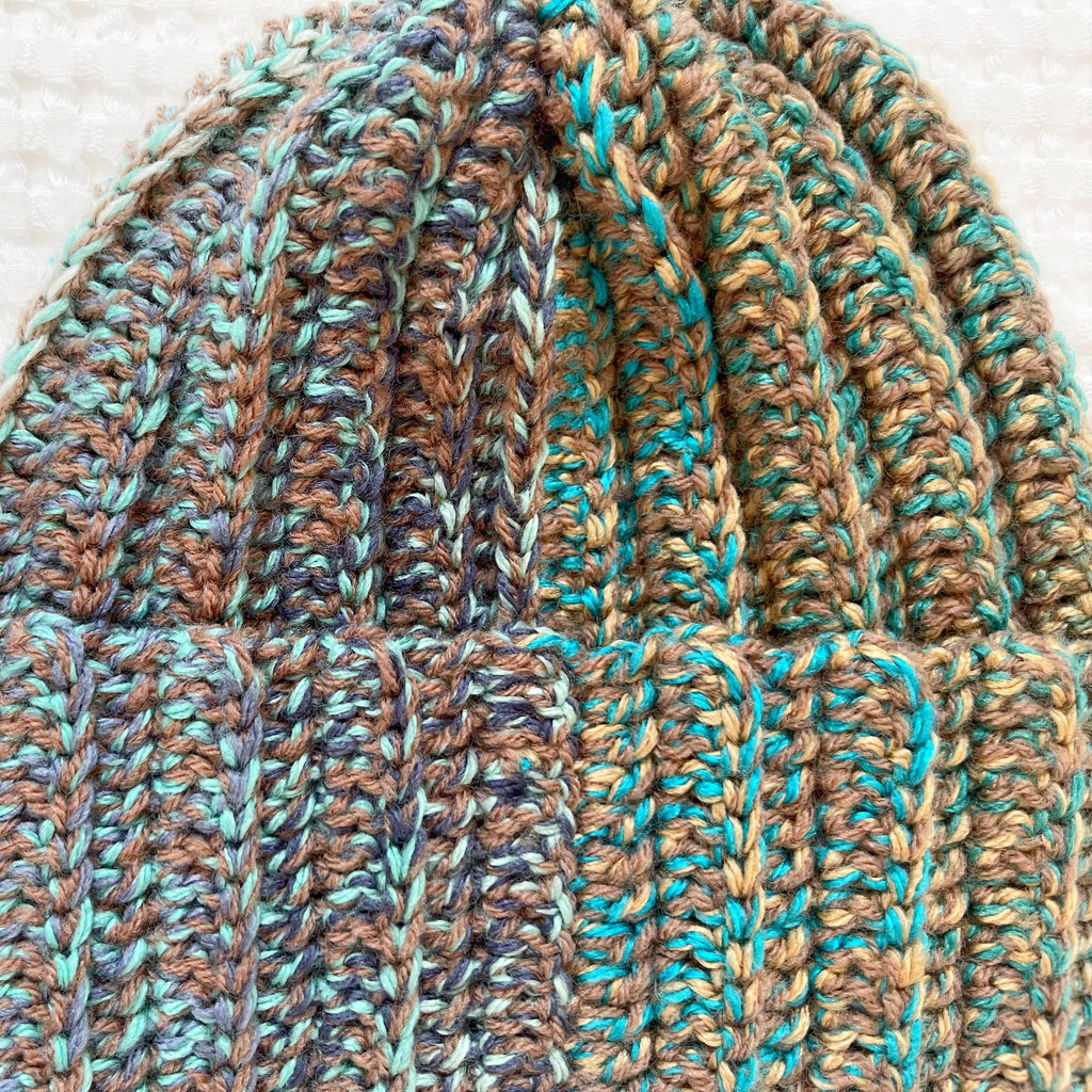 Close-up of a crochet beanie with brown and teal colors on a textured white background